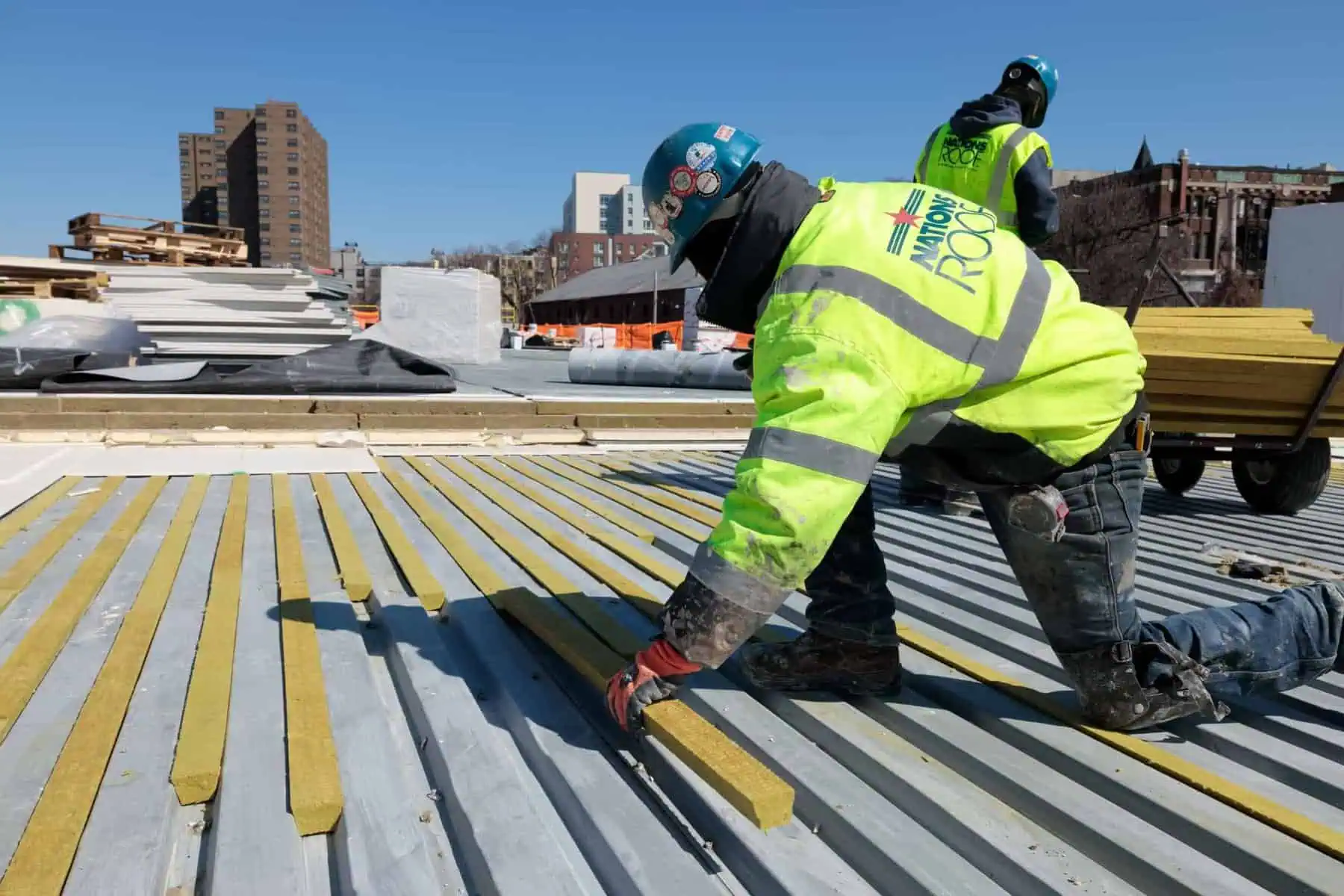 Roofers installing yellow insulation on commercial rooftop. Helmed in green safety gear.