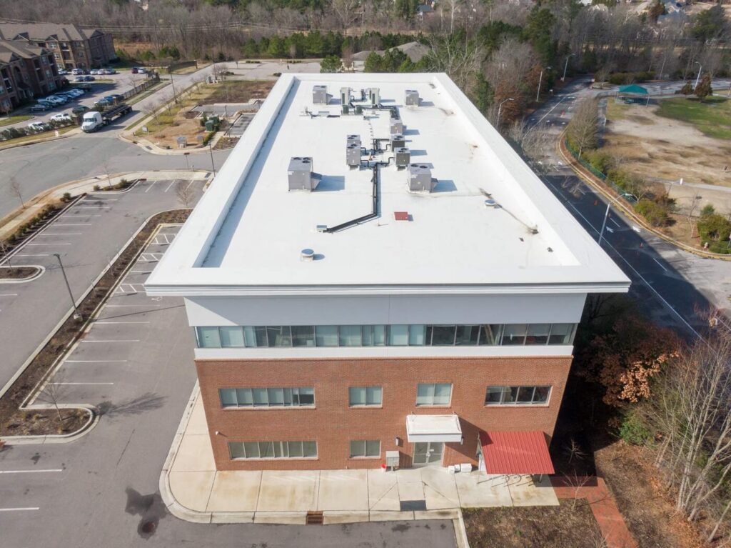 Aerial view of a commercial building with flat white roof and HVAC units. Brick facade, parking lot, and surrounding area visible.