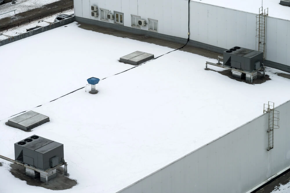 Multi-level commercial flat roof with air handlers and skylights covered in snow.