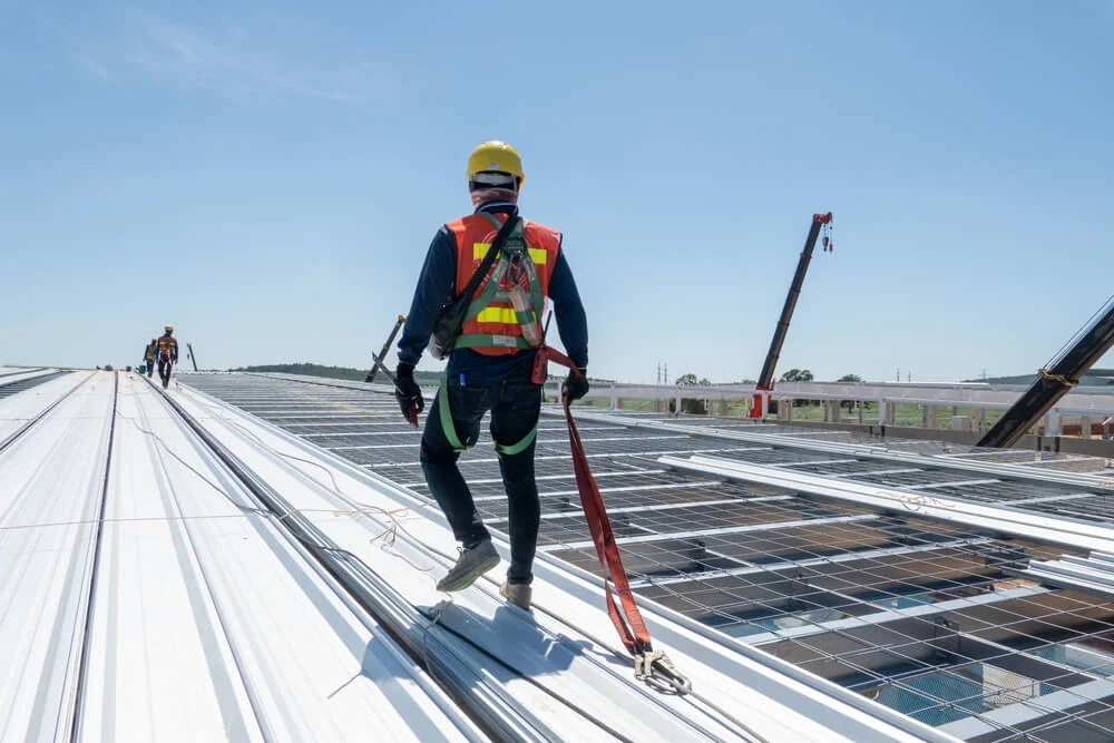 A worker in safety gear on a flat roof with solar panels, secured by a safety line, exemplifies commercial roof maintenance and repair.